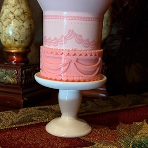 May include: A pink and white decorative cake stand. The stand features a two-tiered design with heart-shaped accents and a scalloped edge. The base is white and the cake stand is made of ceramic.