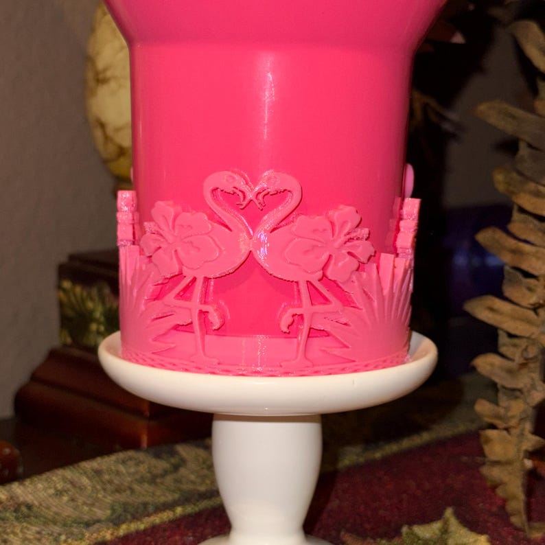May include: A bright pink cylindrical container featuring a raised relief design of two flamingos facing each other, surrounded by hibiscus flowers and palm leaves. The container is supported by a smooth, white, pedestal-style base.
