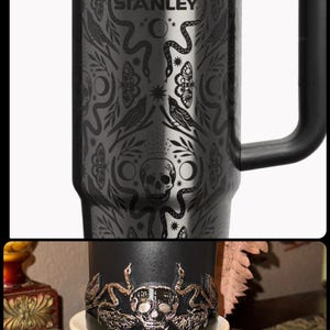 May include: Black Stanley travel mug with a black handle, featuring a detailed engraved design of snakes, butterflies, moons, and a skull. The Stanley logo is visible. The bottom image shows a black candle with a skull and snake design.