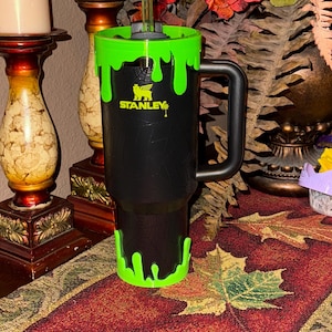May include: A black Stanley travel mug with a green slime design. The mug has a black handle and a black lid with a straw hole. The mug is sitting on a brown and green patterned surface.