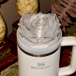 May include: White travel mug with a silver glitter rose on top. The mug has a black Stanley logo on the side.