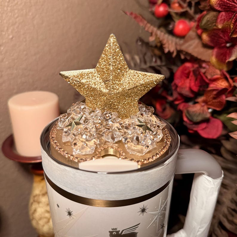 May include: A white and gold mug with a gold glitter star on top. The star is surrounded by clear crystals. The mug has a handle and a decorative design with stars and a logo. The mug is likely for beverages.