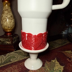 May include: A white tumbler featuring a prominent red plastic band around its lower section. The red band displays a detailed, raised, repeating heart-shaped pattern with intricate textures. The tumbler has a smooth white finish and a handle.