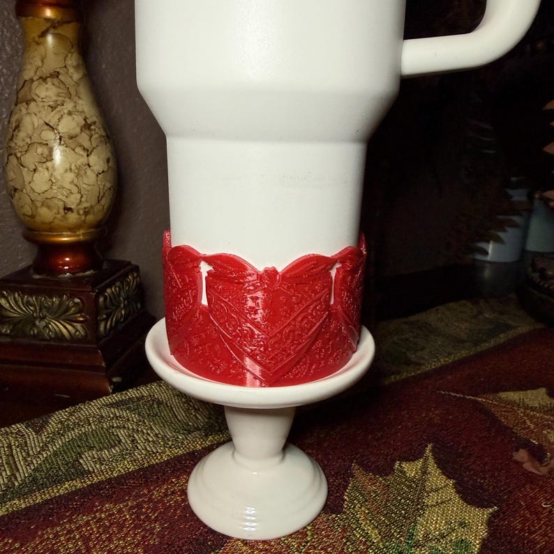 May include: A white tumbler featuring a prominent red plastic band around its lower section. The red band displays a detailed, raised, repeating heart-shaped pattern with intricate textures. The tumbler has a smooth white finish and a handle.