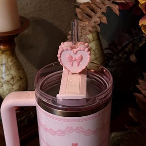 May include: A pink insulated travel mug with a matching handle and lid. The mug has a decorative pink bow and ribbon design. A pink heart-shaped straw topper with a bow detail sits on the lid. A clear straw is inserted into the lid.