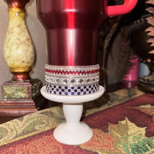 May include: A red insulated tumbler with a pink handle, adorned with three decorative bands. The bands feature red, silver, and blue rhinestones, resting on a white pedestal. The tumbler is displayed on a table with a patterned tablecloth.