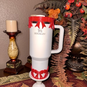 May include: A white Stanley travel mug with a handle, featuring a red bow design around the top and bottom. The Stanley logo is visible on the front. The mug is placed on a white stand, with a candle and fall foliage in the background.