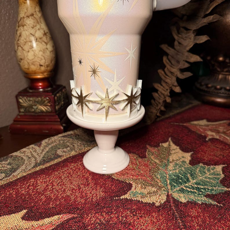 May include: A light pink pedestal candle holder with a white base and a gold star-shaped crown. The candle holder supports a white and gold tumbler with a starburst design. The tumbler is decorated with gold and black stars.