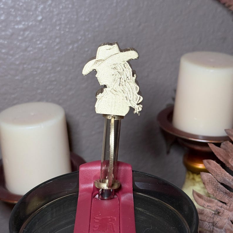 May include: A gold-colored bottle stopper featuring a silhouette of a woman wearing a cowboy hat. The stopper is attached to a burgundy-colored band and is set against a blurred background with candles.