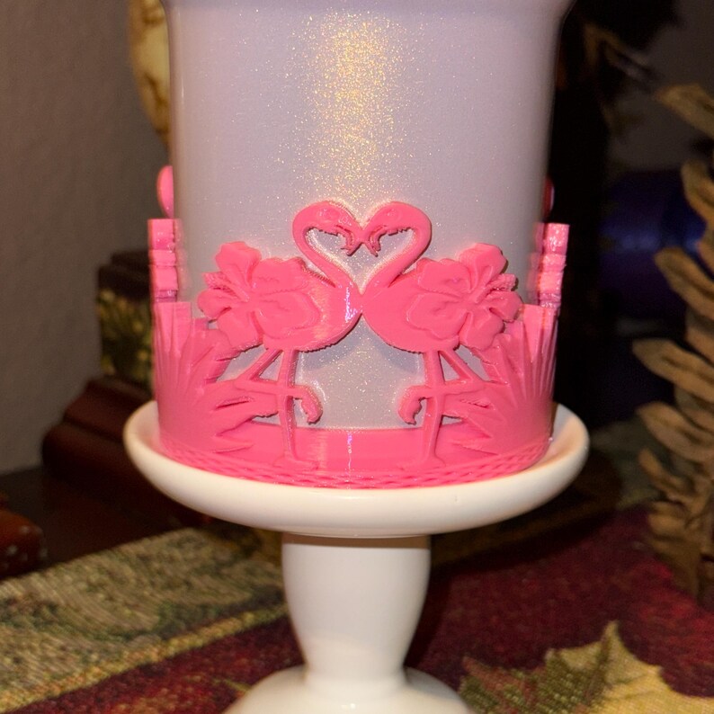 May include: A white cylindrical candle holder with a shimmering finish, featuring a bright pink wrap-around overlay. The overlay displays two flamingos facing each other with their necks forming a heart shape, surrounded by hibiscus flowers and leaves.