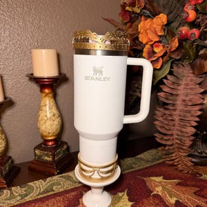 May include: A white Stanley travel mug with a handle, adorned with a gold-colored crown-like embellishment at the top and a gold bracelet at the base. The mug has the Stanley logo and is set on a white pedestal.