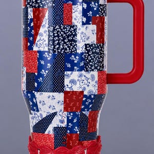 May include: A tall travel mug featuring a patchwork pattern of red, white, and blue squares with various floral, paisley, and polka dot prints. The mug has a solid red lid, a red handle, and a decorative red base with an embossed floral design.
