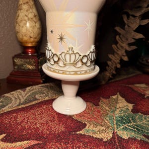 May include: A light pink pedestal candle holder with a gold crown design around the middle. The candle holder has a pearlescent finish with starburst and star designs. The base is a light pink colour.