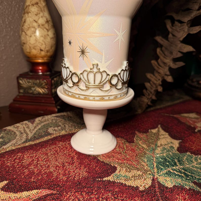 May include: A light pink pedestal candle holder with a gold crown design around the middle. The candle holder has a pearlescent finish with starburst and star designs. The base is a light pink colour.