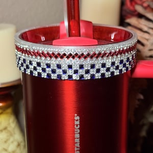 May include: A red Starbucks tumbler with a decorative band of red, white, and blue rhinestones. The tumbler has a red lid and straw. The Starbucks logo is visible on the side of the tumbler.