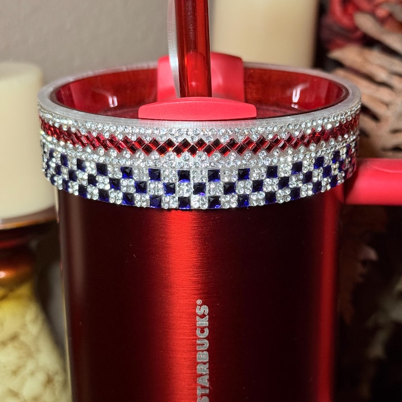 May include: A red Starbucks tumbler with a decorative band of red, white, and blue rhinestones. The tumbler has a red lid and straw. The Starbucks logo is visible on the side of the tumbler.