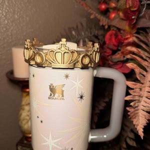 May include: A white Stanley mug with a gold crown lid. The mug features a light pink and white design with star and snowflake patterns. The Stanley logo is visible on the front.