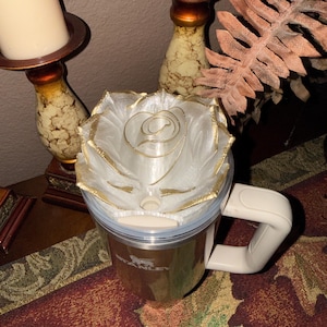 May include: A stainless steel travel mug with a white handle and a decorative rose-shaped lid. The lid is white with gold accents, and the mug has the brand name "Stanley" printed on it. The mug is sitting on a patterned surface.