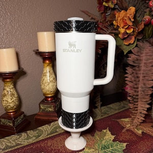 May include: A white Stanley travel mug with a black textured lid and base, featuring the Stanley logo. The mug has a white handle and is displayed on a white pedestal. The background includes candles and fall foliage.