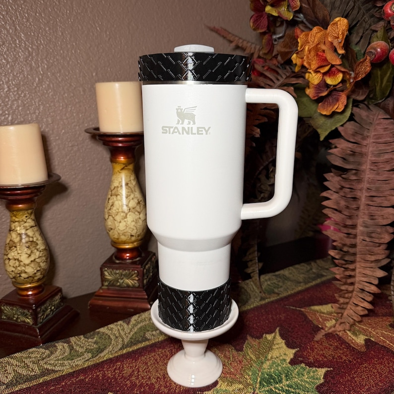 May include: A white Stanley travel mug with a black textured lid and base, featuring the Stanley logo. The mug has a white handle and is displayed on a white pedestal. The background includes candles and fall foliage.
