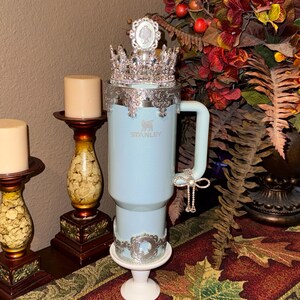 May include: A light blue Stanley tumbler featuring a silver crown topper, a silver filigree rim, a silver bow charm attached to the handle, and a silver filigree band around the base. The tumbler has a smooth finish and the Stanley logo is visible.