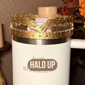 May include: A white Stanley tumbler with a gold-colored decorative crown featuring heart and butterfly cutouts. The text "HALO UP" is printed on the tumbler. A candle and fall foliage are in the background.
