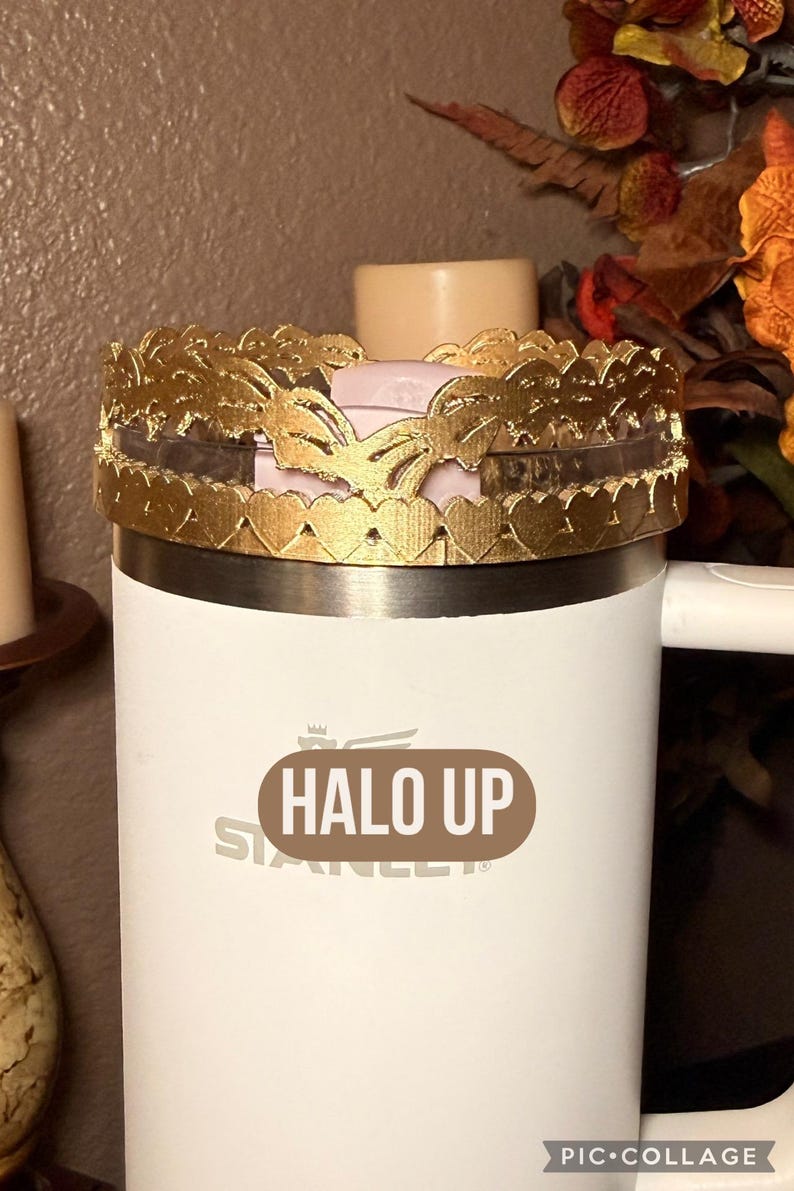 May include: A white Stanley tumbler with a gold-colored decorative crown featuring heart and butterfly cutouts. The text "HALO UP" is printed on the tumbler. A candle and fall foliage are in the background.