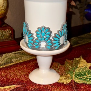 May include: A white candle holder with a turquoise and gray floral design around the base. The candle holder sits on a white pedestal base. The candle holder is on a table with a red and gold patterned tablecloth.