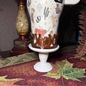 K&ouml;nnte beinhalten: Ein wei&szlig;er Becher mit einem Western-Thema-Design mit Kakteen, Stiefeln und Langhorn-Rindern. Der Becher steht auf einem wei&szlig;en Sockel mit einem braunen hufeisenf&ouml;rmigen Halter. Der Becher ist mit "40oz" beschriftet.