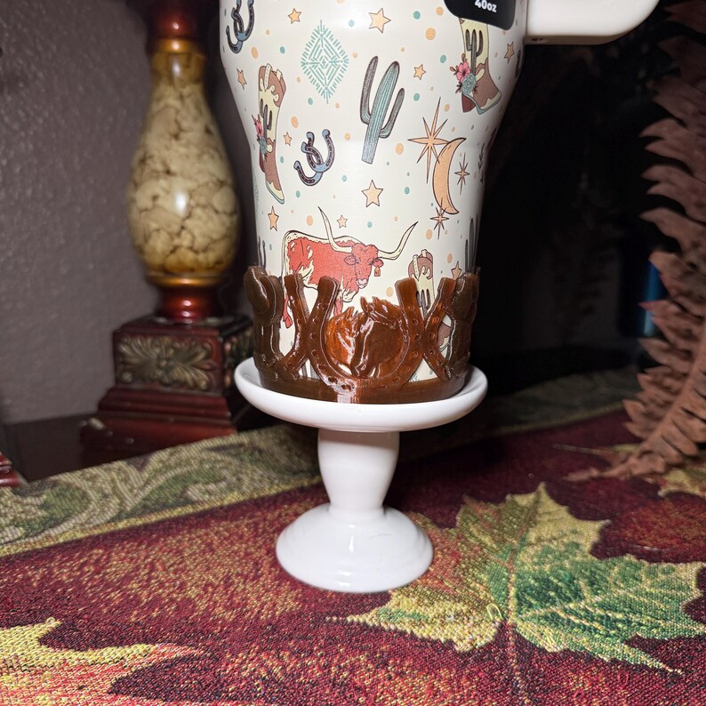 K&ouml;nnte beinhalten: Ein wei&szlig;er Becher mit einem Western-Thema-Design mit Kakteen, Stiefeln und Langhorn-Rindern. Der Becher steht auf einem wei&szlig;en Sockel mit einem braunen hufeisenf&ouml;rmigen Halter. Der Becher ist mit "40oz" beschriftet.