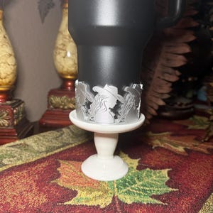 May include: A black tumbler with a decorative silver band featuring a silhouette of a woman wearing a cowboy hat. The tumbler sits on a white pedestal base, creating a unique and stylish presentation.