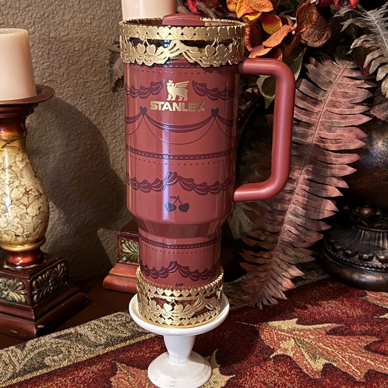 May include: A maroon Stanley travel mug with a gold filigree design around the top and bottom. The mug has a handle and a lid. The Stanley logo is visible. The mug is decorated with a repeating pattern of cherries and decorative elements.