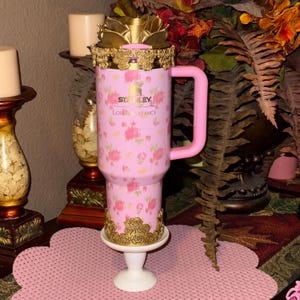 May include: A pink Stanley tumbler with a floral pattern and the words "Love & Fancy." The tumbler has a pink handle and is decorated with gold accents. It is placed on a white pedestal.