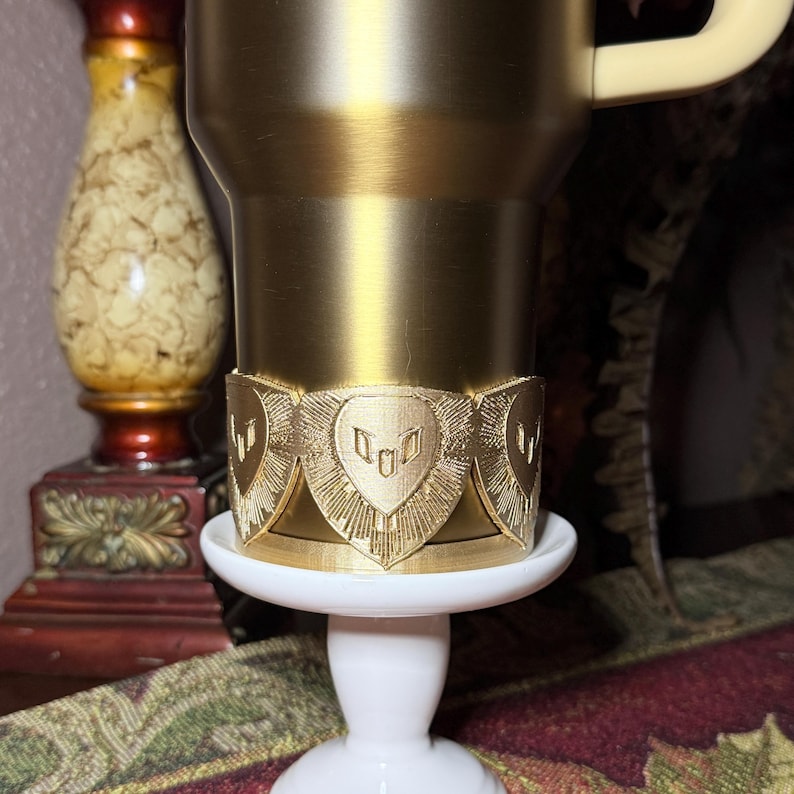 May include: A gold-colored travel mug with a cream-colored handle, resting on a white pedestal. The mug features a decorative gold band with a repeating shield design. The overall aesthetic is ornate and elegant.