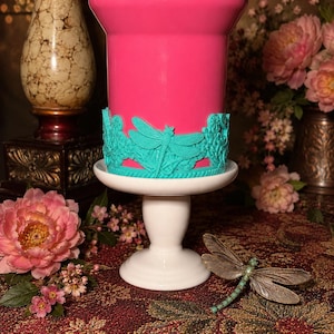 May include: A bright pink cylindrical container sits on a white pedestal stand. The base of the container is wrapped in a teal decorative band featuring a raised dragonfly and floral relief pattern. A separate dragonfly ornament rests nearby.