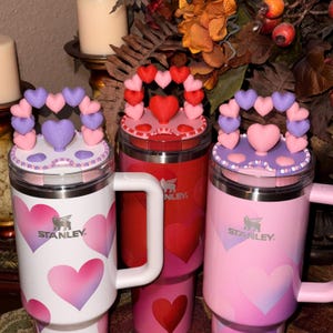 May include: Three Stanley travel mugs with heart designs. One is white with pink hearts, another is red, and the third is pink. Each mug has a heart-shaped topper with a heart-shaped handle. The Stanley logo is visible.