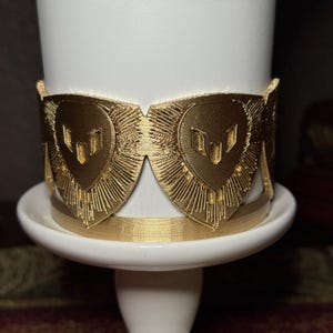 May include: A gold-colored decorative band with a heart-shaped design, featuring a series of vertical lines radiating outwards. The band is wrapped around a white cylindrical object, resting on a white base.