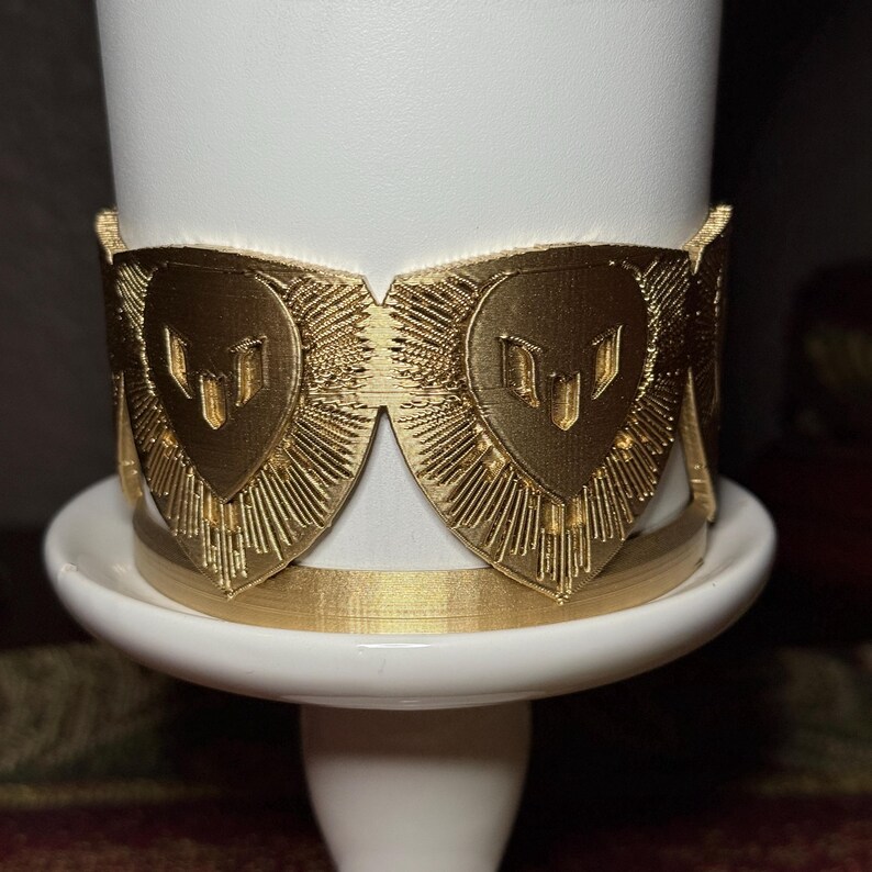 May include: A gold-colored decorative band with a heart-shaped design, featuring a series of vertical lines radiating outwards. The band is wrapped around a white cylindrical object, resting on a white base.