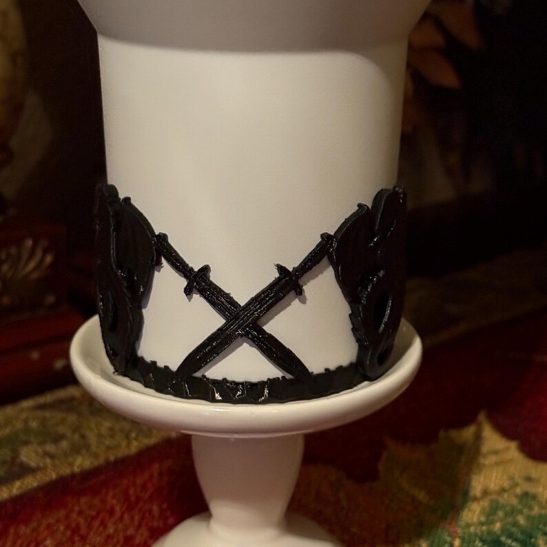 May include: White ceramic candle holder with a black 3D printed design of crossed swords and floral accents.