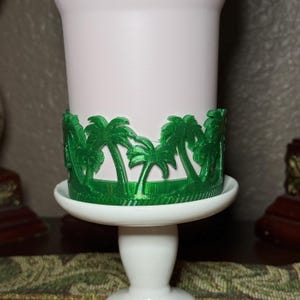 May include: A white cylindrical cup holder featuring a vibrant green, three-dimensional wrap-around design of palm trees. The cup holder sits securely on a white pedestal base, showcasing a detailed, textured botanical pattern in a bright green finish.