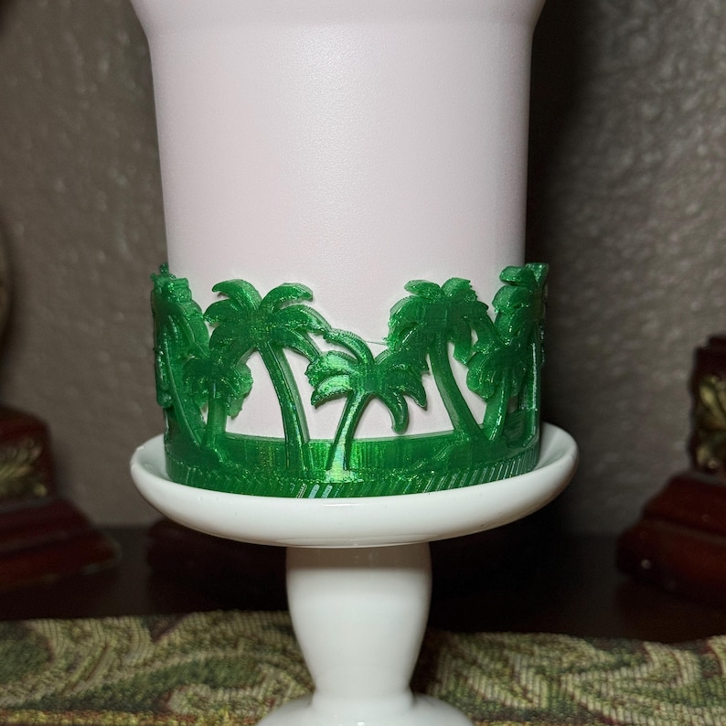 May include: A white cylindrical cup holder featuring a vibrant green, three-dimensional wrap-around design of palm trees. The cup holder sits securely on a white pedestal base, showcasing a detailed, textured botanical pattern in a bright green finish.