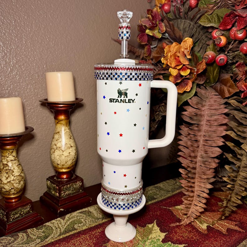 May include: White Stanley travel mug with a handle, decorated with red, blue, and black stars. The mug has a decorative rim and base with rhinestones. A clear, jeweled topper is on the lid. The Stanley logo is visible.