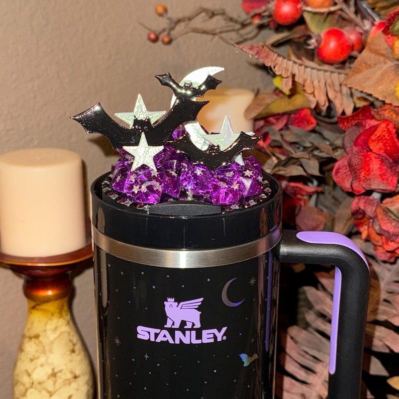 May include: Black Stanley travel mug with a purple handle and accents. The mug features the Stanley logo and a crescent moon design. Topped with purple candies and Halloween-themed bat and star decorations.