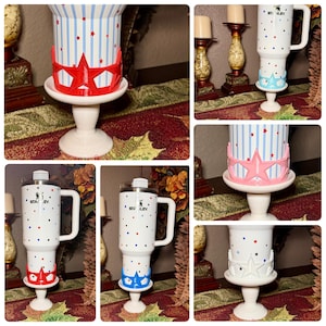 May include: A white Stanley tumbler with a handle, decorated with red, blue, and white stars and stripes. The tumbler sits on a white pedestal base with a decorative star-shaped accent in red, pink, blue, or white. The tumbler has a clear lid.