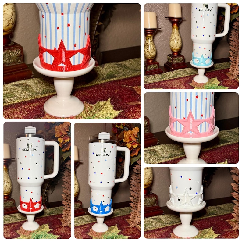 May include: A white Stanley tumbler with a handle, decorated with red, blue, and white stars and stripes. The tumbler sits on a white pedestal base with a decorative star-shaped accent in red, pink, blue, or white. The tumbler has a clear lid.