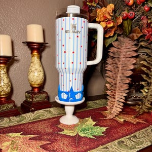 May include: A stainless steel Stanley tumbler with a white handle, adorned with blue vertical stripes and red star accents. The tumbler sits on a white stand with a blue star-shaped design. The Stanley logo is visible.