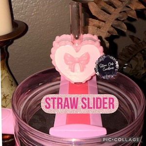 May include: Pink straw slider featuring a heart-shaped design with a bow. The text "STRAW SLIDER" is displayed on the slider. The slider is designed to fit on a tumbler or cup.