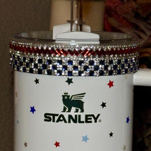 May include: A white Stanley tumbler with a decorative rim featuring red, white, and blue rhinestones. The tumbler has a black logo and star accents in red, blue, and black. The tumbler has a clear lid and a handle.