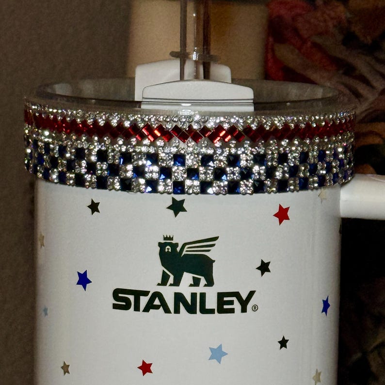 May include: A white Stanley tumbler with a decorative rim featuring red, white, and blue rhinestones. The tumbler has a black logo and star accents in red, blue, and black. The tumbler has a clear lid and a handle.