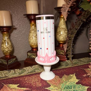 May include: A Stanley brand tumbler with a pink and white striped pattern, accented with red and blue stars. The tumbler sits on a white pedestal with a pink star-shaped base. The tumbler has a silver top.